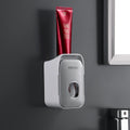 Automatic Toothpaste Squeezer in light gray plastic with a rounded wall-mounted dispenser holding a red toothpaste tube