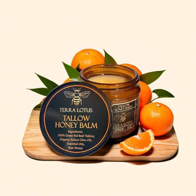 Honey Tallow Moisturizing Balm in an amber glass jar with a black-and-gold label, alongside fresh oranges and leaves.